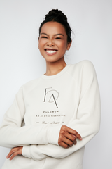 Model wearing a cream Fulcrum crewneck sweater with minimalist black logo, smiling softly.