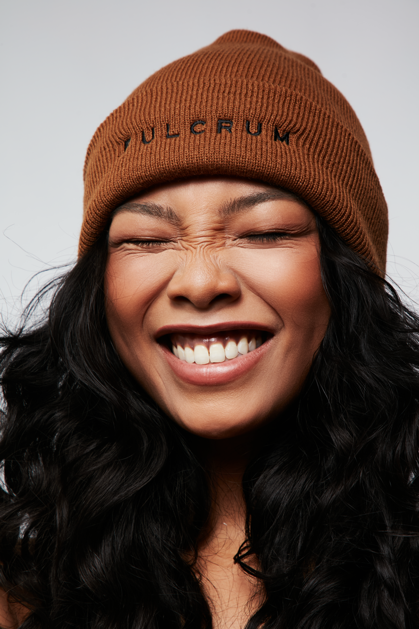 Close-up of model wearing a brown ribbed Fulcrum beanie with black embroidered logo, smiling with eyes closed.